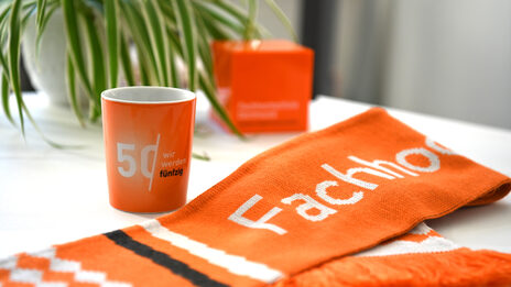 A cup and a scarf are lying on a table. Both are orange. The scarf says "Fachhochschule" and the mug says "Wir werden fünfzig".