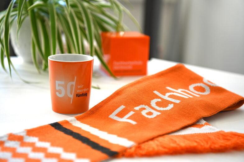 A cup and a scarf are lying on a table. Both are orange. The scarf says "Fachhochschule" and the mug says "Wir werden fünfzig".