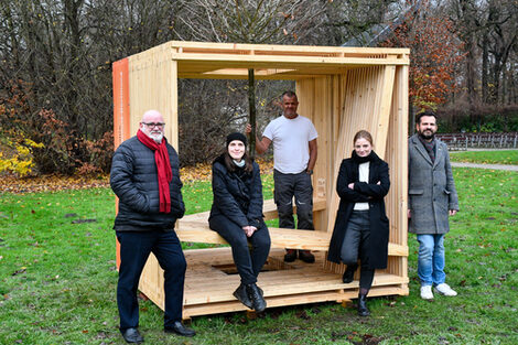 A group of people have gathered around a wooden cube in a park.