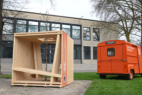 Eine würfelartige Holzkonstruktion steht neben einem orangefarbenen Citroen HY.