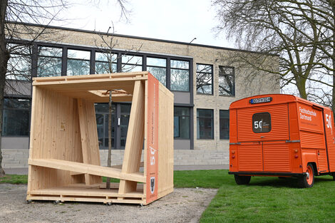 A cube-like wooden structure stands next to an orange Citroen HY.