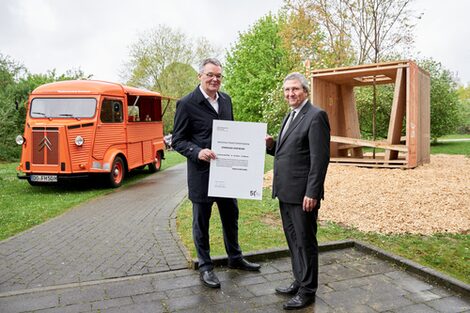 Two male figures stand in front of the wooden construction of a three-dimensional cube. They are holding a certificate in their hands.