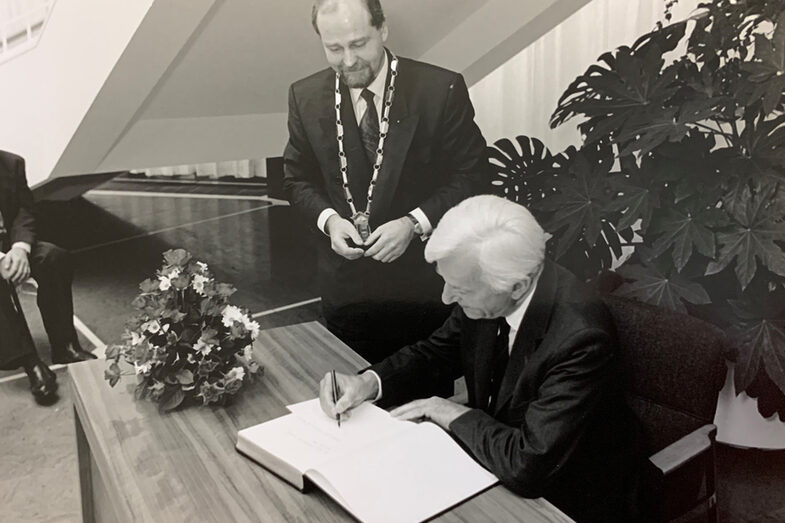 A man is sitting at a table writing in a book. A second man with a chain of office stands next to him and watches.