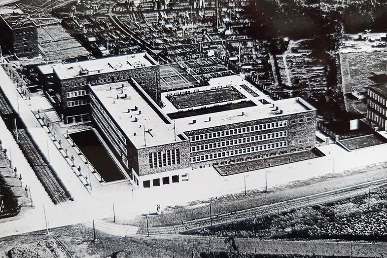 Das Schwarz-Weiß-Foto zeigt einen großen Gebäude-Komplex. Rechts davon ist Industrie zu sehen, die Bundesstraße 1 im Vordergrund ist nur eine schmale Straße.