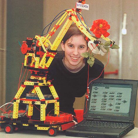 A young woman next to a kind of toy crane with a gripper arm. Next to it is a laptop. The robotic arm is holding a rose.