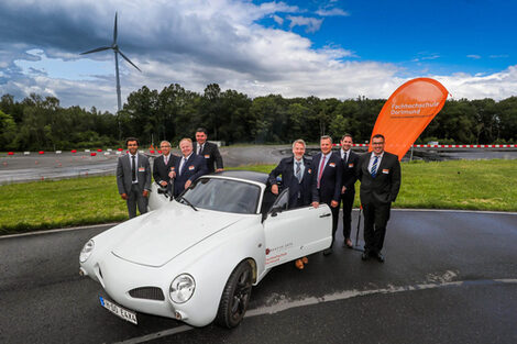 Photo from the opening ceremony of the cooperation between Fachhochschule Dortmund and Lasise. Eight men in suits stand around a car with the FH lettering on the door. An FH flag in the background.