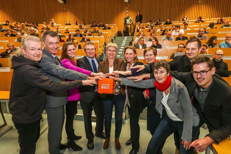 Eine Gruppe von Menschen steht in einem Hörsaal. Alle haben eine Hand auf einen orangefarbenen Würfel gelegt.