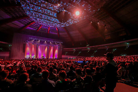 Many people are sitting in the illuminated Westfalenhalle. They are looking at a stage.