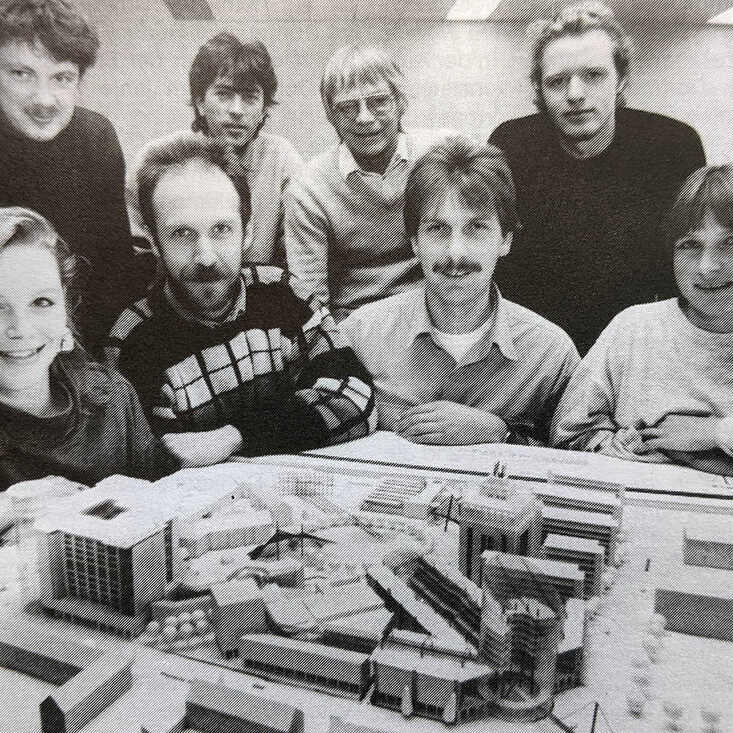 A group of people squat around an architectural model and look into the camera. The Dortmunder U is clearly visible on the model.