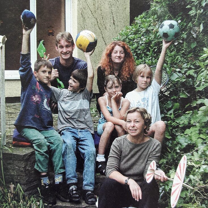 Four children and three adults are sitting on an outside staircase in front of a building. The children are holding up balls.