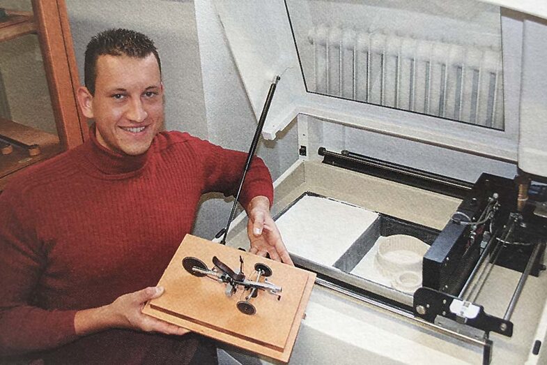 A young man holds a printed bicycle model in his hand. He is sitting in front of a 3D printer.