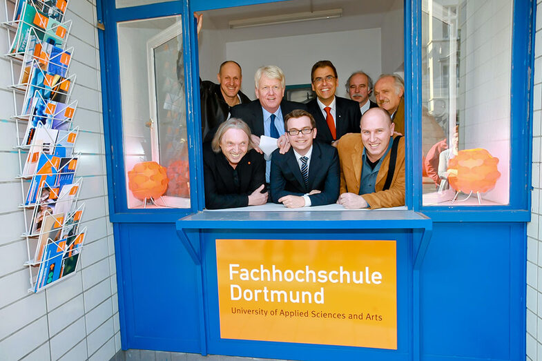 A group of men peer out of a blue kiosk window. A sign reading "Fachhochschule Dortmund" hangs below the window.