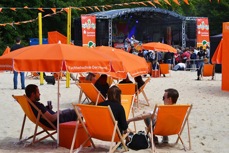 People sit on orange sun chairs under orange parasols. A stage can be seen in the background.