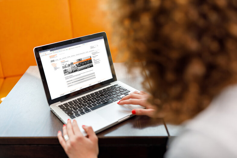 Foto einer jungen am Laptop, im Hintergrund sind orangefarbene Sitzmöbel. __ Young woman at the laptop, in the background there are orange colored chairs.