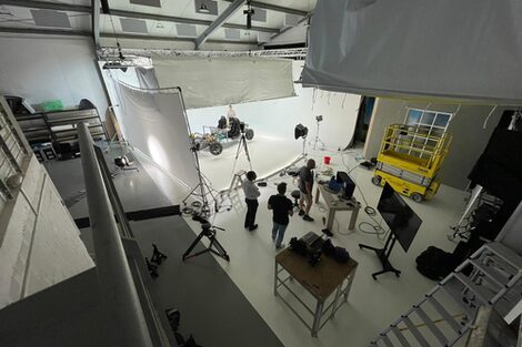 From above, you look into a room where a photo shoot is taking place. In an area cordoned off by sheets, you can see the vehicle being photographed. In the foreground are three people and various pieces of equipment and devices.