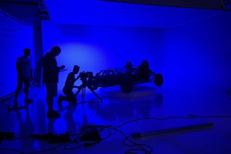 In a dark, blue-lit room, you can see the black outline of a car body in the middle of the room. To the left, a man is kneeling in front of a video camera. Two other men are standing next to him. You can only make out the black outlines of them too.