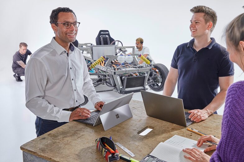 Three people are standing at a table in the foreground: an older man on the left at his laptop, smiling. To his right is a young man, also at his laptop, who is looking at the other man. From diagonally behind, you can also see a young woman looking at a notepad in front of her. In the background, two young men are working on a car body.