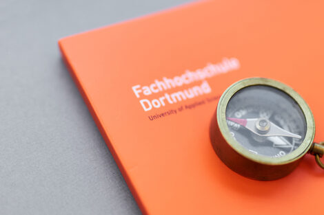 Foto eines kleinen Kompasses mit Kette daran, der auf einer orangefarbenen Mappe der Fachhochschule Dortmund liegt.