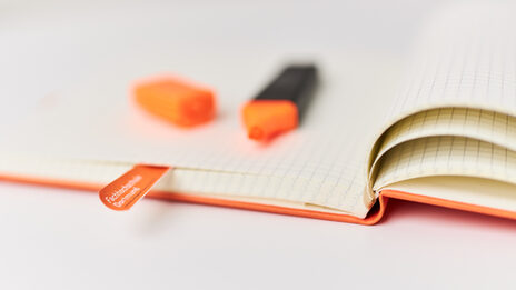 Nahaufnahme eines Notizbuches, auf dem ein geöffneter orangefarbener Textmarker liegt. Ein Zettel mit Aufdruck „Fachhochschule Dortmund“ markiert eine Seite.