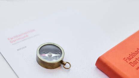 Photo of various things on a table: a notepad from Fachhochschule Dortmund, with a small compass on it and part of a notebook next to it.