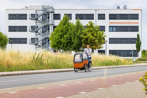 Foto von einem Mann auf einem Lastenfahrrad, der auf der Straße von einem Gebäude wegfährt. Auf dem Gebäude ist der Schriftzug "Fachhochschule Dortmund" zu sehen.
