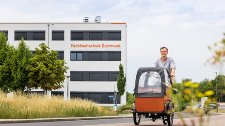 Foto von einem Mann auf einem Lastenfahrrad, der auf dem Radweg von einem Gebäude wegfährt. Auf dem Gebäude ist der Schriftzug "Fachhochschule Dortmund" zu sehen.