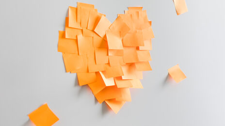 Foto von einem aus orangenen Haftnotizzetteln zusammengeklebten Herz an einer Wand. __ A heart stuck together from orange sticky notes on the wall.