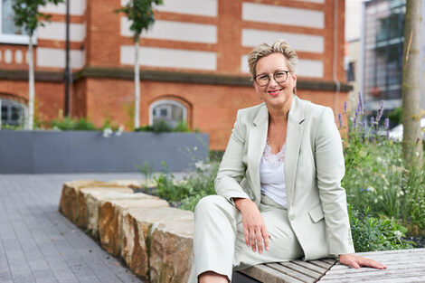 Eine Frau in einem grünen Blazer sitzt draußen vor einem Gebäude und lacht.__A woman in a green blazer sits outside in front of a building and laughs