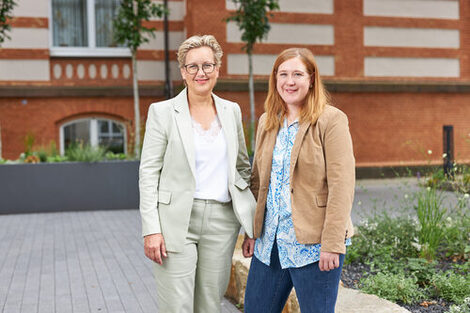 Zwei Frauen mit Blazer stehen vor einem Gebäude und lächeln id die Kamera.__Two women in blazers stand in front of a building and smile for the camera