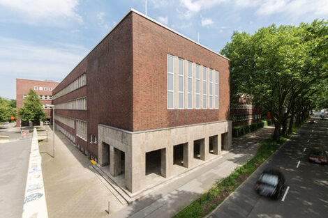 Photo with a view of parts of a building of the Fachhochschule Dortmund at Max-Ophüls-Platz with forecourt and sidewalk as well as street with cars next to it.