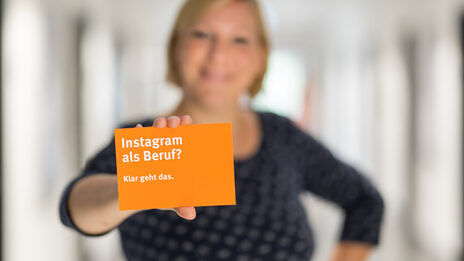 Foto von einer Frau - unscharf zu erkennen - die eine orangefarbene Postkarte der Fachhochschule in die Kamera hält. Auf der Postkarte steht der Schriftzug "Instagram als Beruf? Klar geht das.".