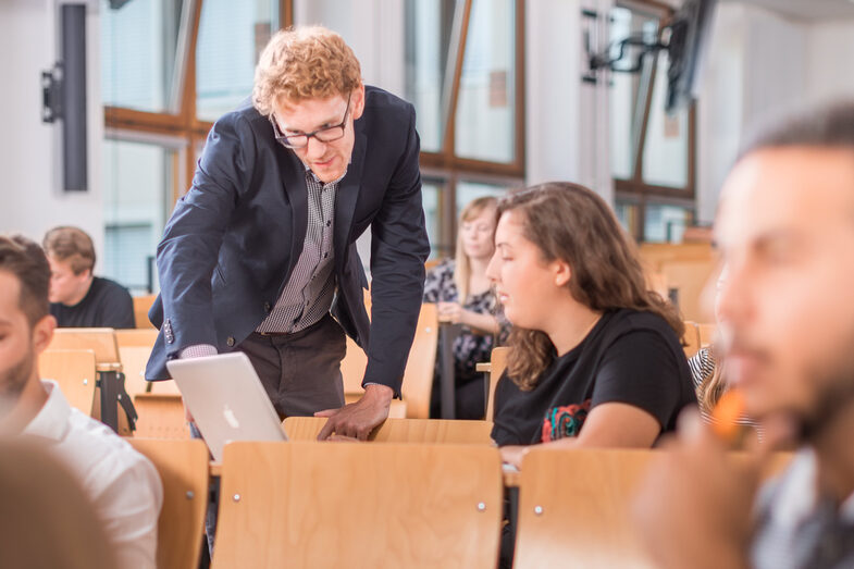 Foto eines Dozenten, der im Hörsaalgang steht und einer Studentin etwas auf ihrem Laptop zeigt. Drumherum unscharf weitere Studierende.__Photo of a lecturer standing in the lecture hall corridor showing a student something on her laptop. Around it blurred other students.