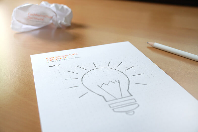 Foto von einer Skizze einer Glühlampe auf einem FH-Block, im Hintergrund zerknülltes Papier und ein FH-Bleistift. __ Sketch of a light bulb on a FH block, crumpled paper in the background and a FH pencil.
