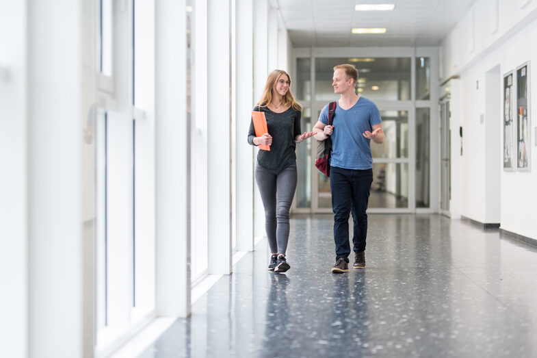 Foto einer Studentin und eines Studenten, die nebeneinander über einen FLur gehen und sich angeregt unterhalten.__One female and one male student walk side by side down the hallway and talk lively.