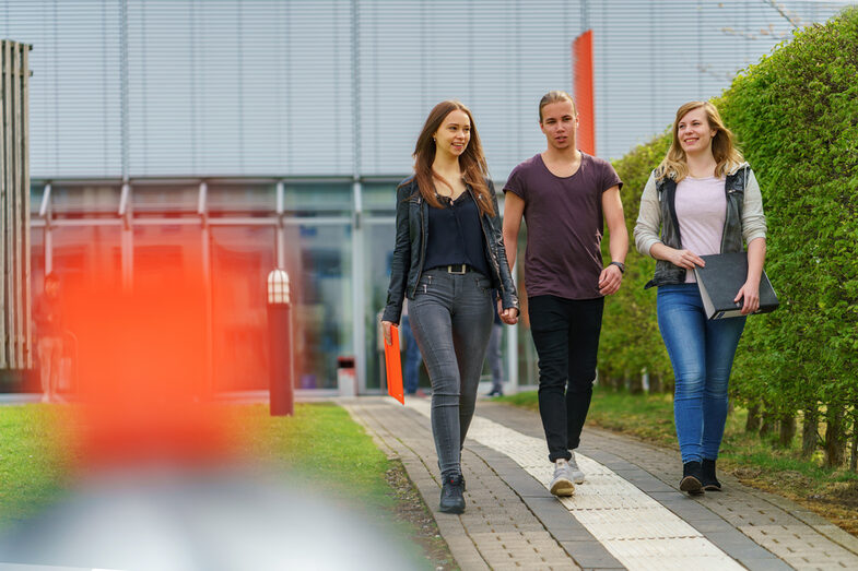 Foto von drei Studierenden, die nebeneinander auf einem Weg von einem Gebäude weg gehen.__Three students walk side by side on a path and move away from a building.