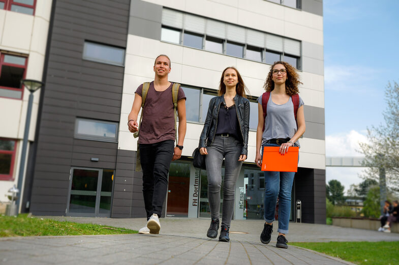 Foto von zwei Studentinnen und einem Studenten, die nebeneinander auf einem Weg gehen, im Hintergrund ein Gebäude in der Emil-Figge-Straße und weitere Studierende.__Two students walk side by side on a path, in the background a building on Emil-Figge-Straße and other students.