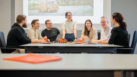 A group of seven people sit around a table while a woman stands looking to the right. A presentation is projected onto the wall in the background. A table with a meeting folder can be seen out of focus in the foreground
