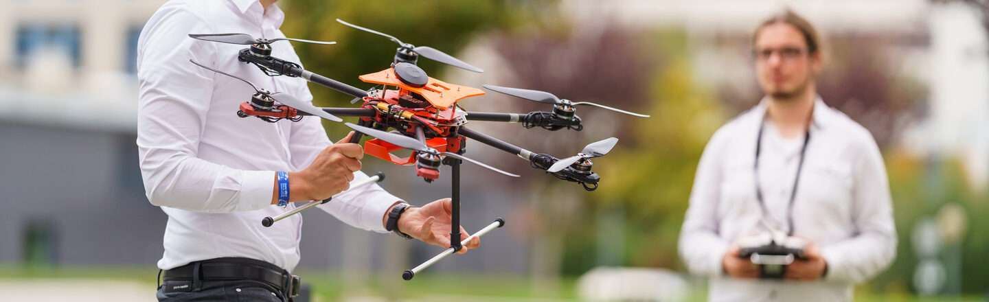 Photo of two IDiAL employees standing opposite each other on a lawn. One of them is holding a drone, the other is holding the controller for controlling the drone.