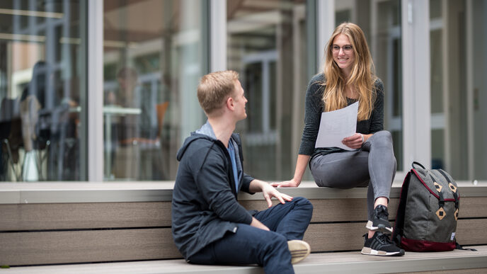 Foto einer Studentin und eines Studenten, die auf dem Campus auf Holzstufen sitzen und sich unterhalten. Die Studentin hält einen Zettel in der Hand.__One male and one female student sitting on wooden steps and talking, the female student is holding a note.