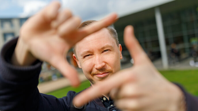 Foto eines Studenten, der mit Zeigefingern und Daumen vor sich ein Quadrat formt und dadurch guckt.
