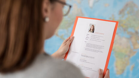 Foto über die Schulter einer Frau. Sie hält eine Mappe mit Bewerbungsunterlagen bzw. einem Lebenslauf in Ihren Händen. Im Hintergrund eine Weltkarte an der Wand. __ Woman holds portfolio with application documents or a résumé in her hands. In the background a world map on the wall.