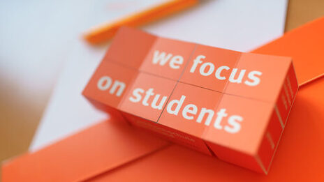 Foto eines aufgeklappten Klappwürfels mit dem Aufdruck "we focus on students", der auf Unterlagen liegt. __ Opened folding cube with the imprint "we focus on students" lies on documents.