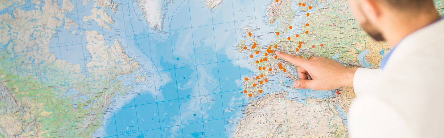 Foto einer Person, die auf eine große Weltkarte zeigt, auf der mit orangefarbenen Punkten Orte markiert sind.