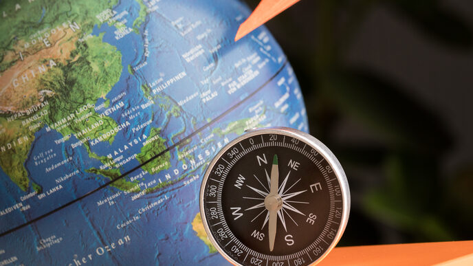 Photo of a still life: globe in the background, FH cube, orange paper airplane and compass in front of it.