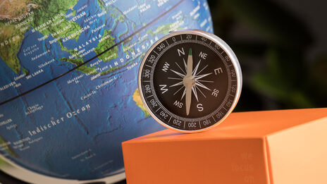 Foto eines Stilllebens: Globus im Hintergrund, davor FH-Würfel, orangener Papierflieger und Kompass.