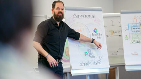 Foto eines Mitarbeiters bei einem Seminarangebot der Personalentwicklung. Er erklärt etwas anhand eines Flipcharts, auf dem Zeichnungen und Beschriftungen zu sehen sind.__Photo of an employee at a seminar offered by the personnel development department. He is explaining something using a flipchart on which drawings and labels can be seen.