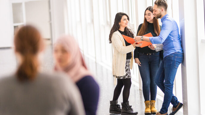 Foto von fünf internationalen Studierenden, von denen sich zwei im Vordergrund unterhalten und die anderen drei gemeinsam in eine orangefarbene Mappe sehen.