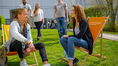 Foto von einer Studentin und einem Studenten in Liegestühlen mit der Aufschrift Fachhochschule Dortmund auf einer Wiese auf dem Campus. Sie unterhalten sich, weitere Studierende nähern sich.__Two students sit in deckchairs with the label Fachhochschule Dortmund on it on the campus and chat. Other students in the background.