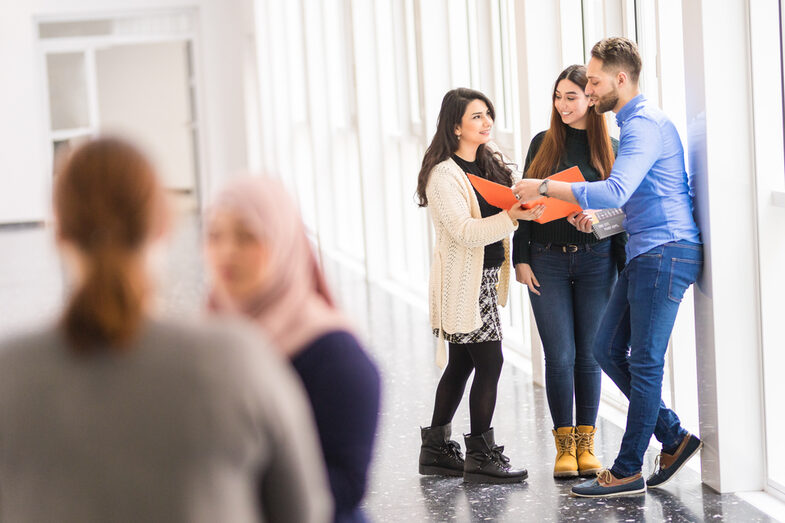 Foto von fünf internationalen Studierenden, von denen sich zwei im Vordergrund unterhalten und die anderen drei gemeinsam in eine orangefarbene Mappe sehen.