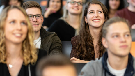 Close-up of several students in the rows of seats in a lecture hall. The focus is on a female student and a male student sitting next to each other and looking straight up.